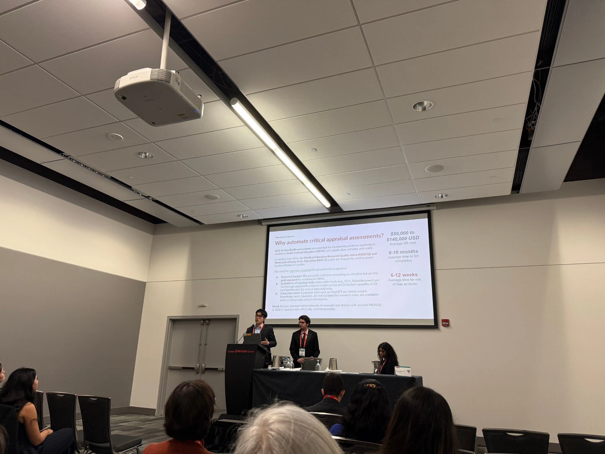 Two Medevi team members on stage presenting the slide "Why automate critical appraisal assessments?" to a seated audience.